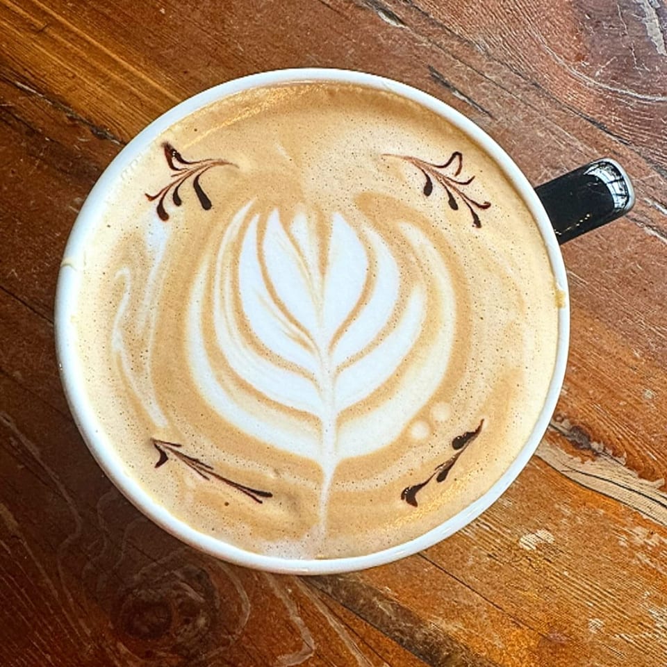Overhead view of a latte with intricate leaf-shaped latte art and chocolate drizzle on a wooden table.