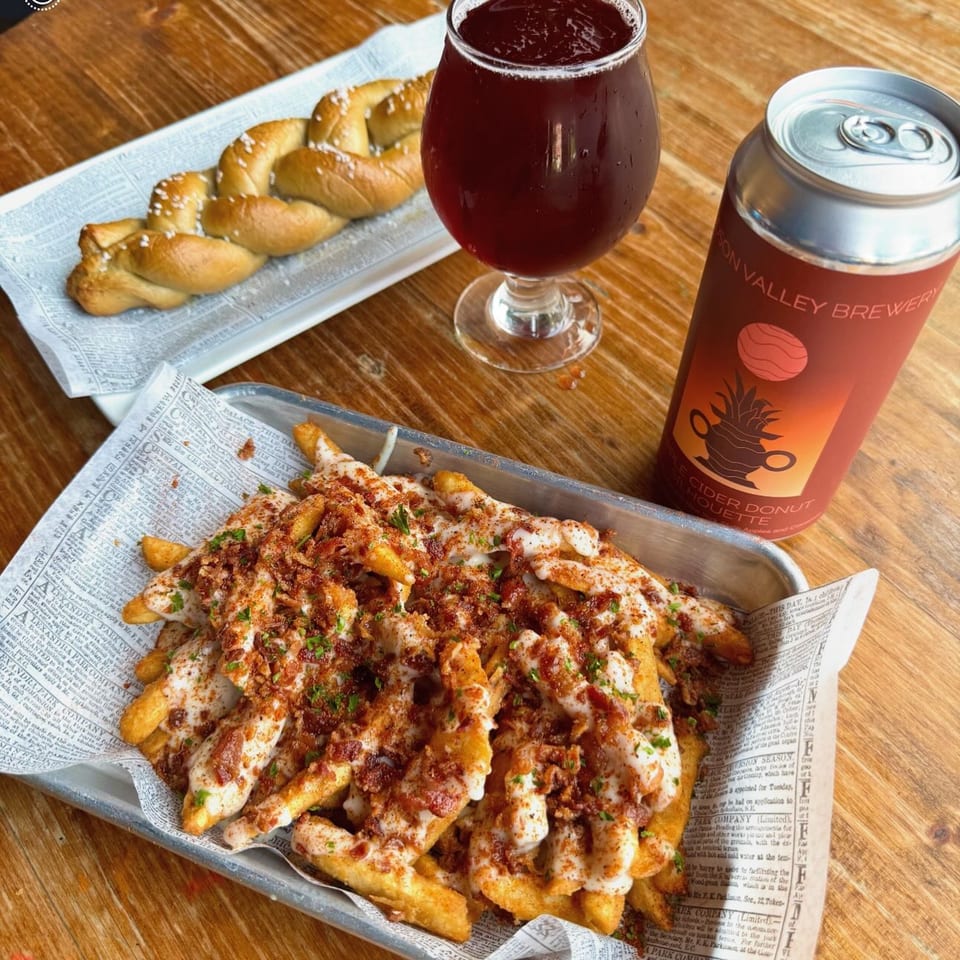 Loaded fries, a braided pretzel, a glass of dark beer, and a beer can on a wooden table.