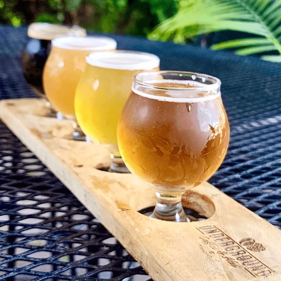 A flight of four craft beers on a wooden paddle, with the Underground Coffee & Ales logo.