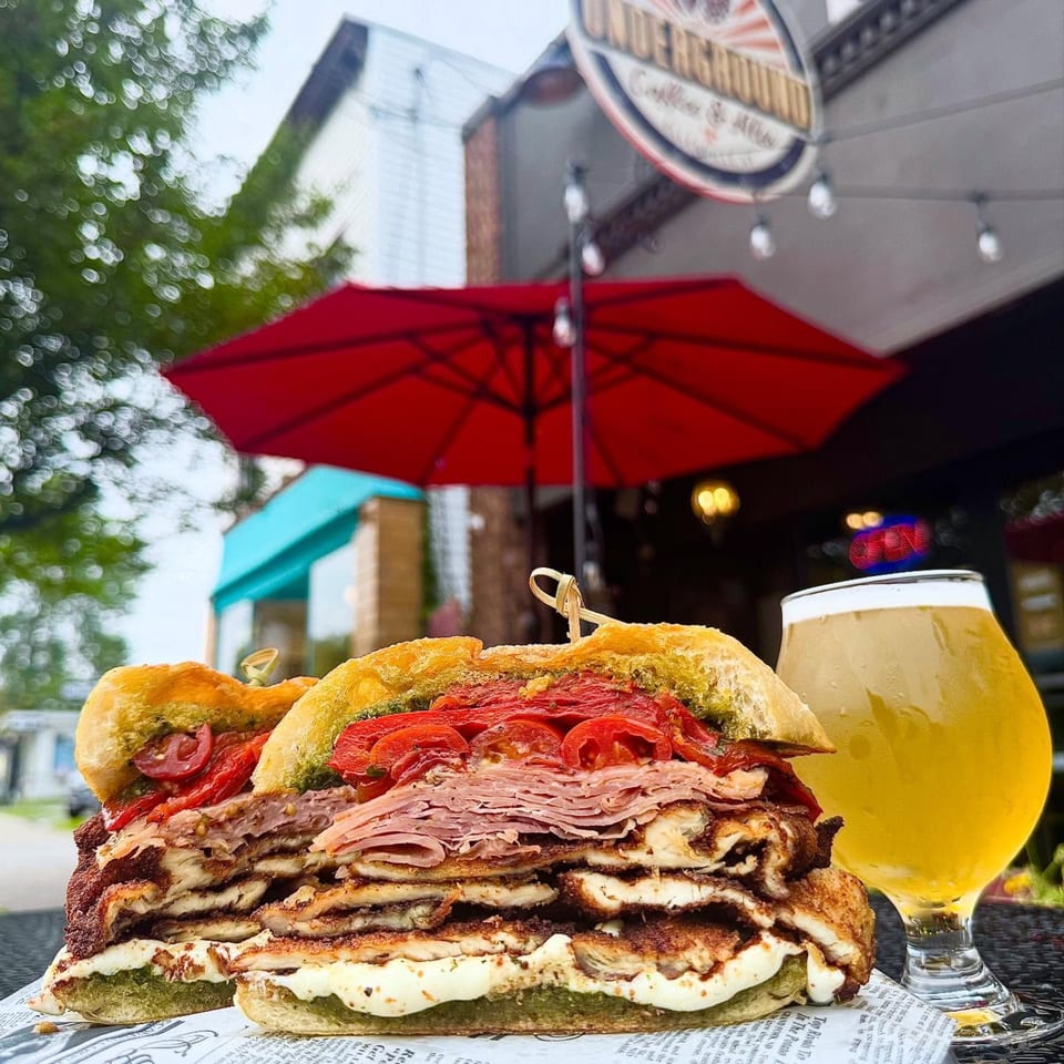 A large sandwich with meat, cheese, and tomatoes, next to a glass of beer outside Underground Coffee & Ales.